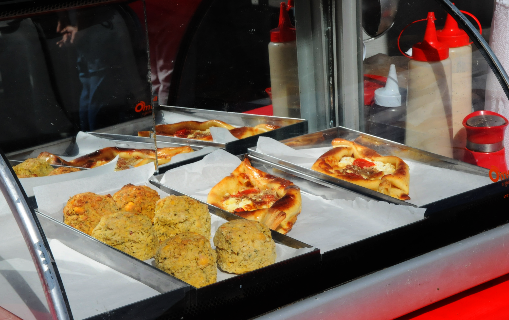 Vitrine de alimentos. É possível ver falafel, um bolinho dourado de gergelim, e esfirras abertas de queijo e tomate.